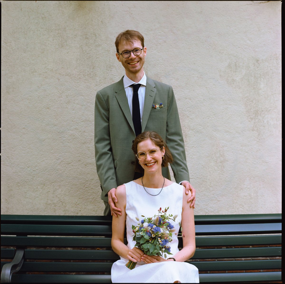 Groom standing behind the bride, who sits on a bench holding her bouquet, medium format film portrait