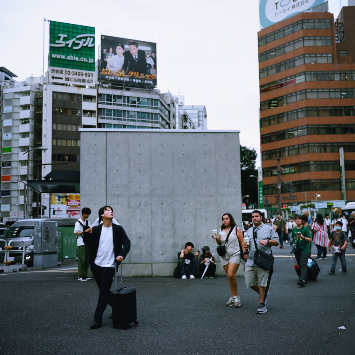Reisender mit Rollkoffer schaut im Gehen nach oben, belebter Platz in Shibuya