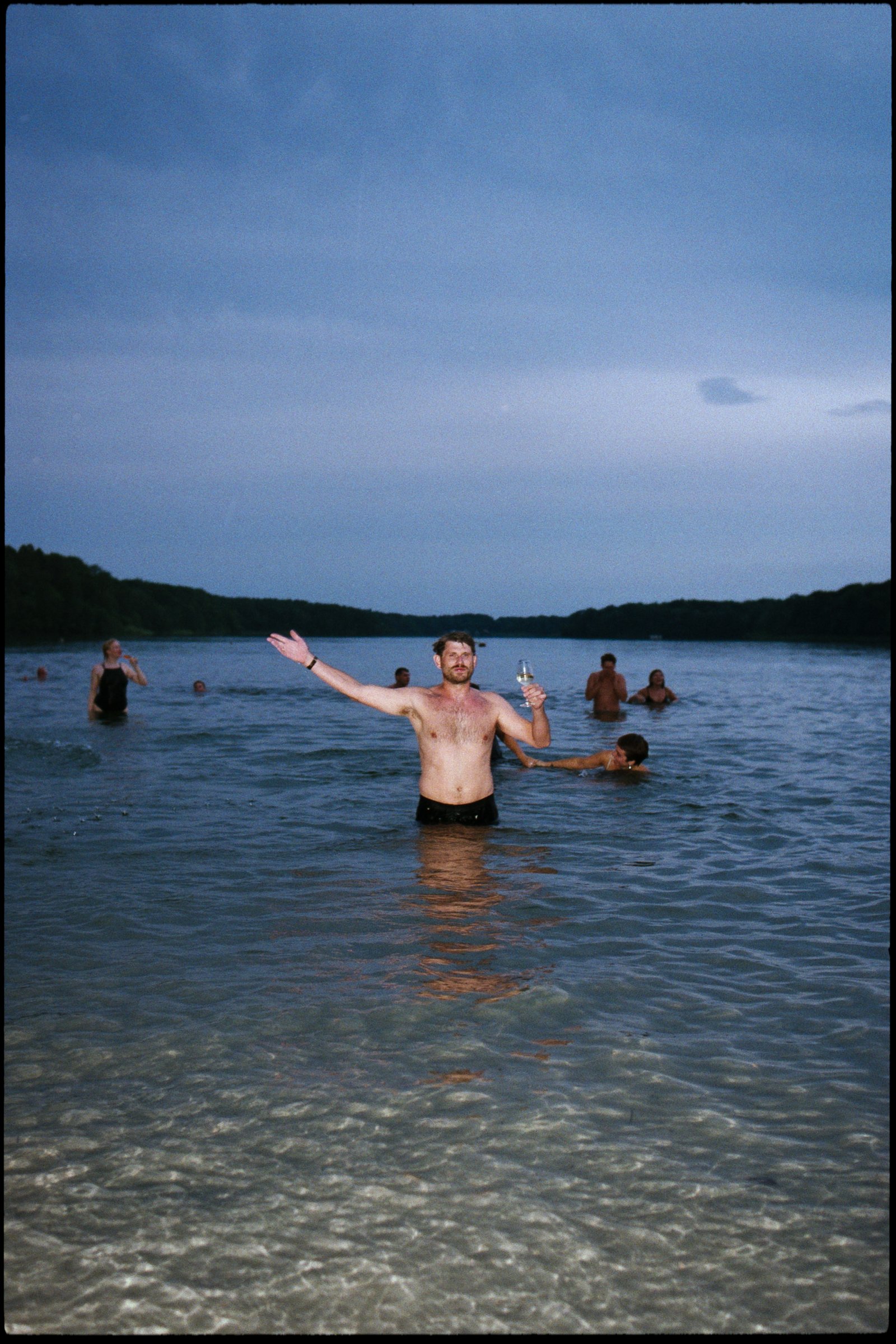 Wedding guest standing waist-deep in a lake at dusk, raising a glass of wine while others swim around him, analogue film photograph